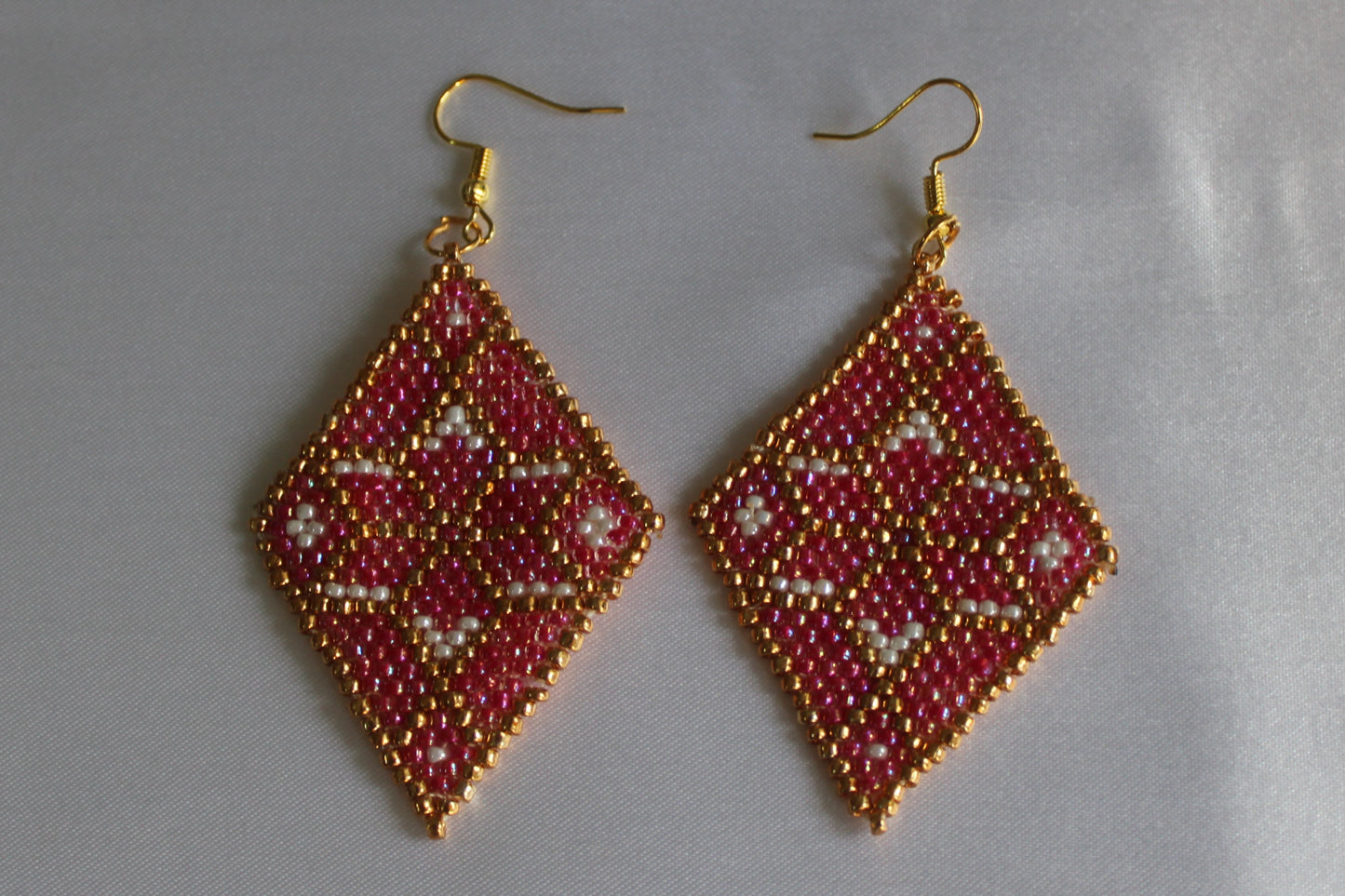 A pair of handmade beaded dangle earrings with gold fishhook wires. The diamond-shaped beads feature a geometric pattern in red, gold, and white, set against a plain light gray background.