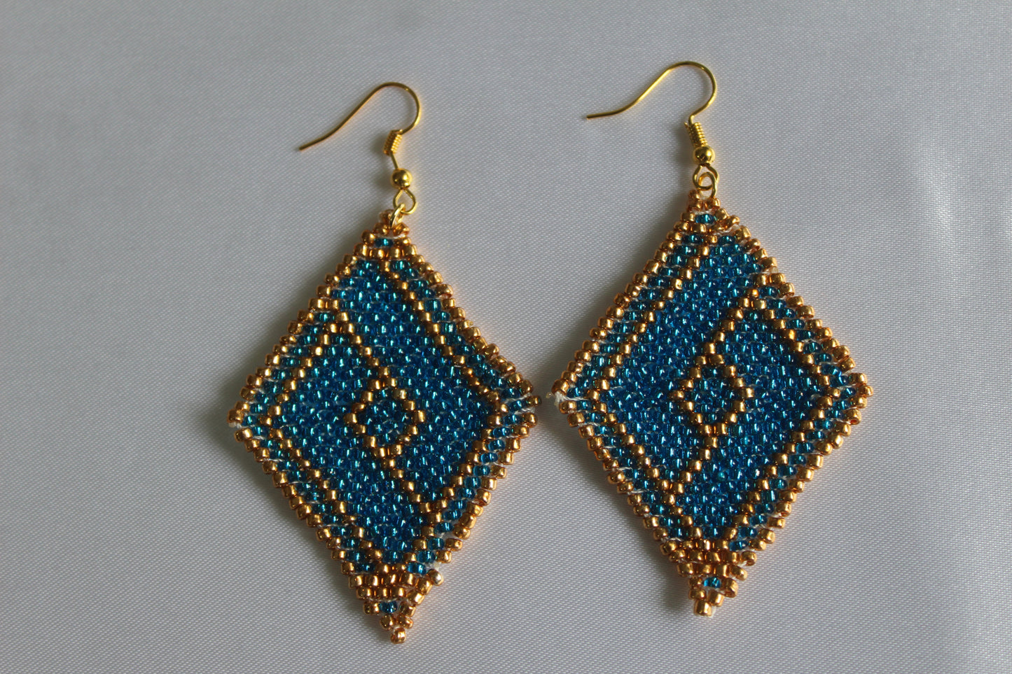 Handmade teal blue and gold diamond-shaped seed bead earrings featuring an intricate geometric pattern with gold-tone fish hook ear wires, lightweight boho statement jewellery.