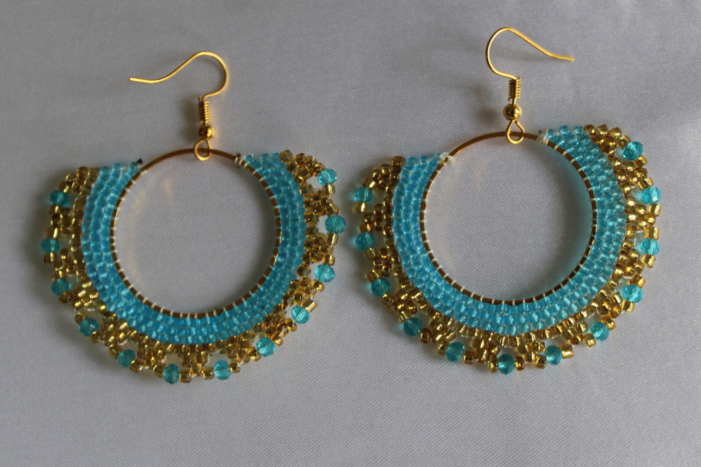Handmade turquoise blue and gold beaded hoop earrings featuring intricate seed bead embroidery on gold-tone hoops with fish hook ear wires.