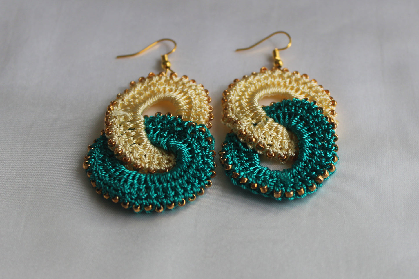 A pair of handmade macrame hoop earrings with a gold-tone metal fishhook and two interlocking spiral loops in teal green and cream, displayed on a light gray fabric background.