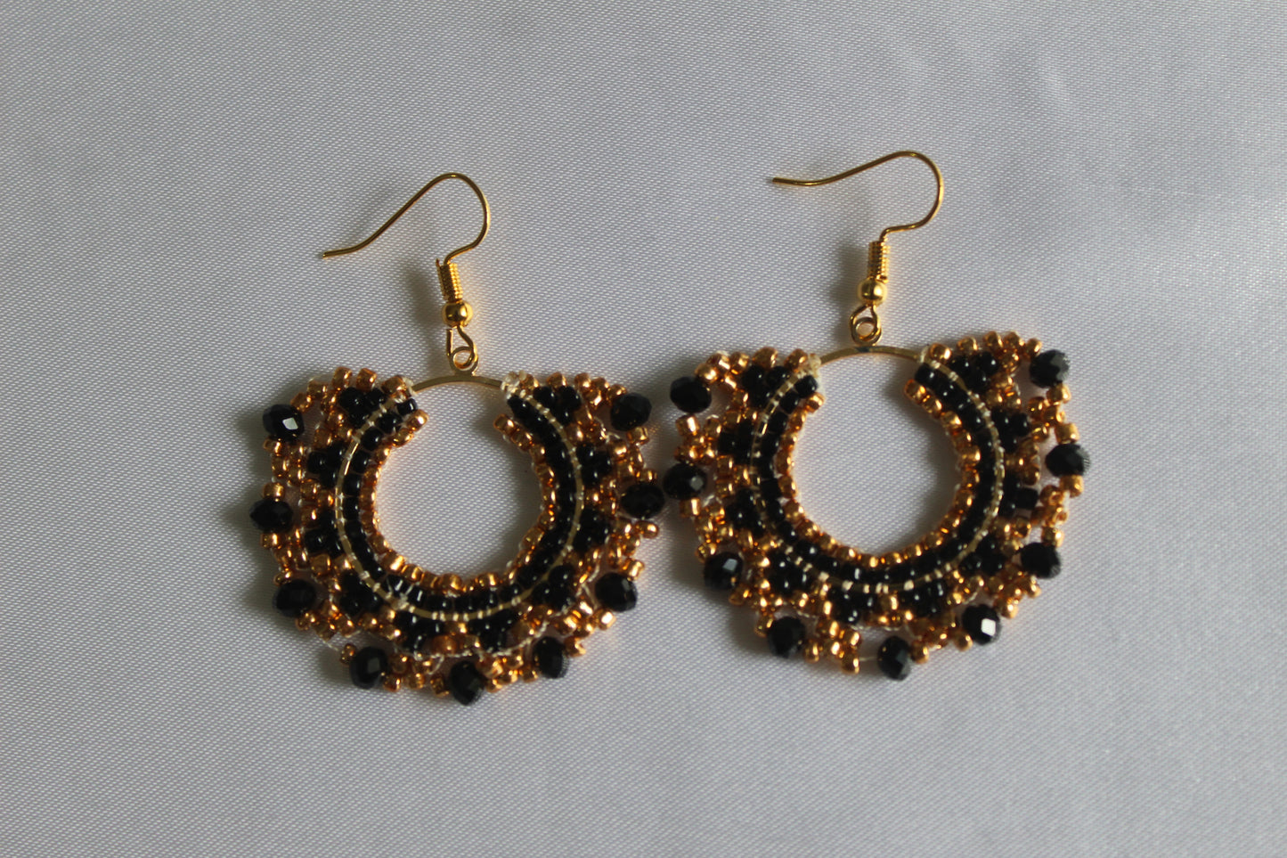 A pair of handmade gold and black beaded hoop earrings with gold fishhook wires, displayed on a light gray fabric background.
