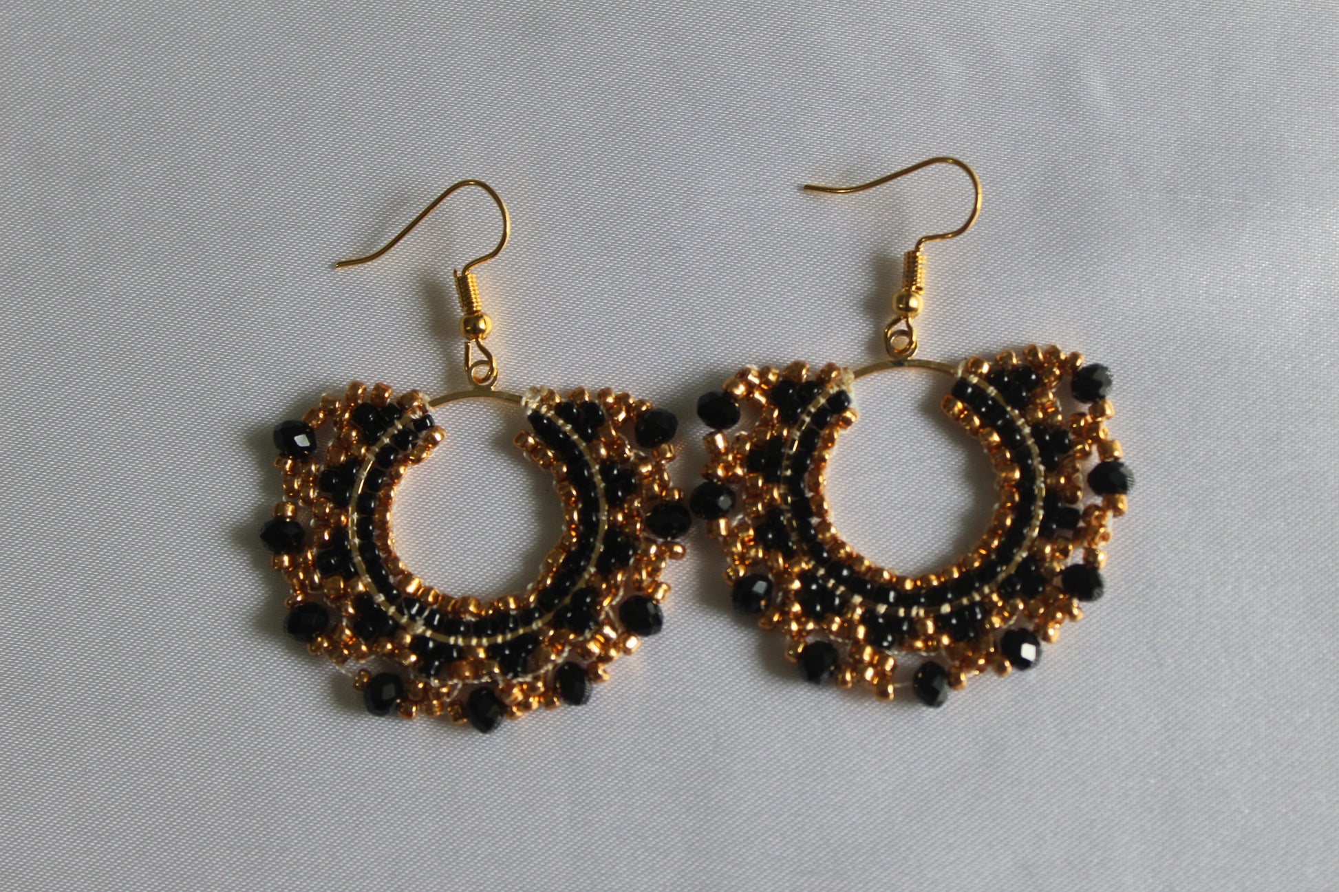 A pair of handmade gold and black beaded hoop earrings with gold fishhook wires, displayed on a light gray fabric background.