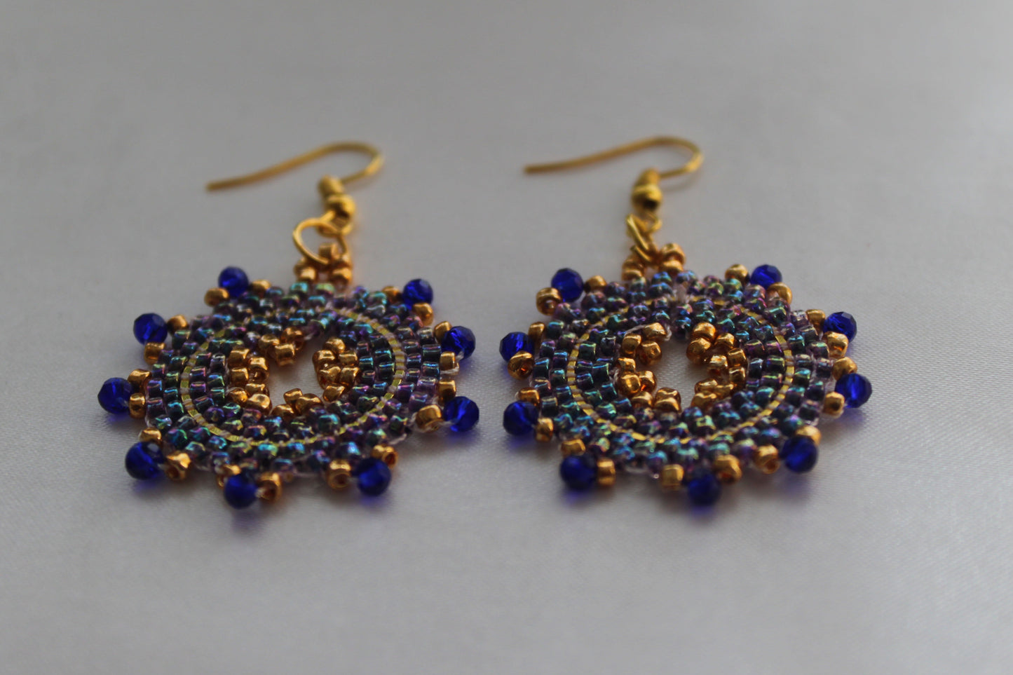 A pair of handmade beaded dangle earrings with gold-tone hooks and circular patterns of dark blue and iridescent blue beads on a light gray background.