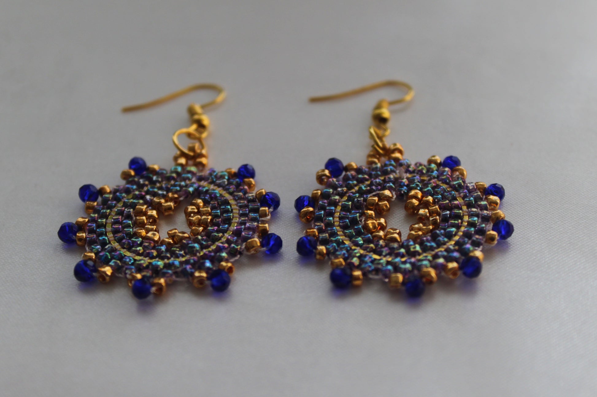 A pair of handmade beaded dangle earrings with gold-tone hooks and circular patterns of dark blue and iridescent blue beads on a light gray background.