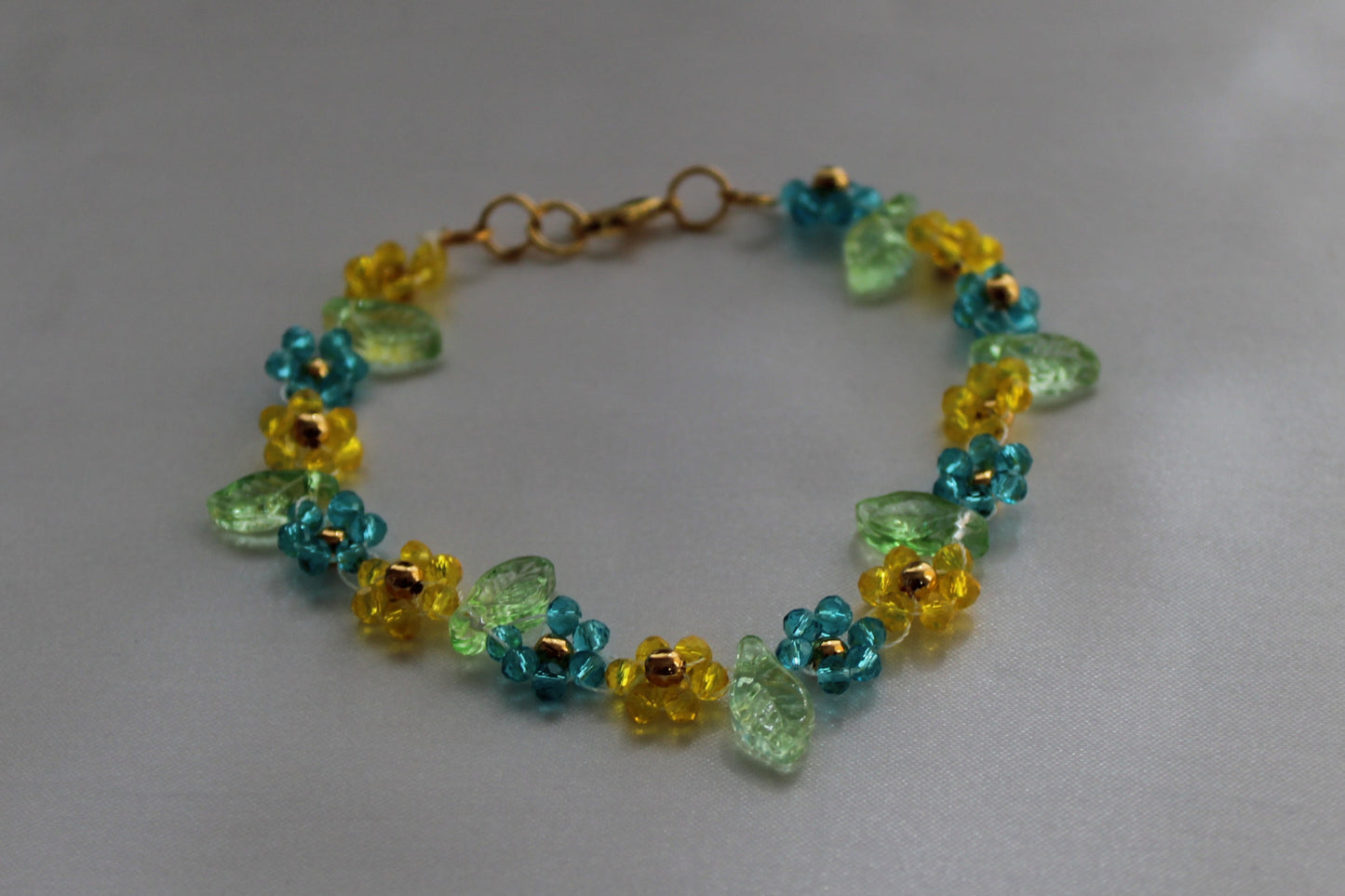 Handmade floral bracelet featuring blue and yellow crystal flowers with translucent green leaf beads and a gold-tone adjustable lobster clasp, lightweight boho handmade jewellery.