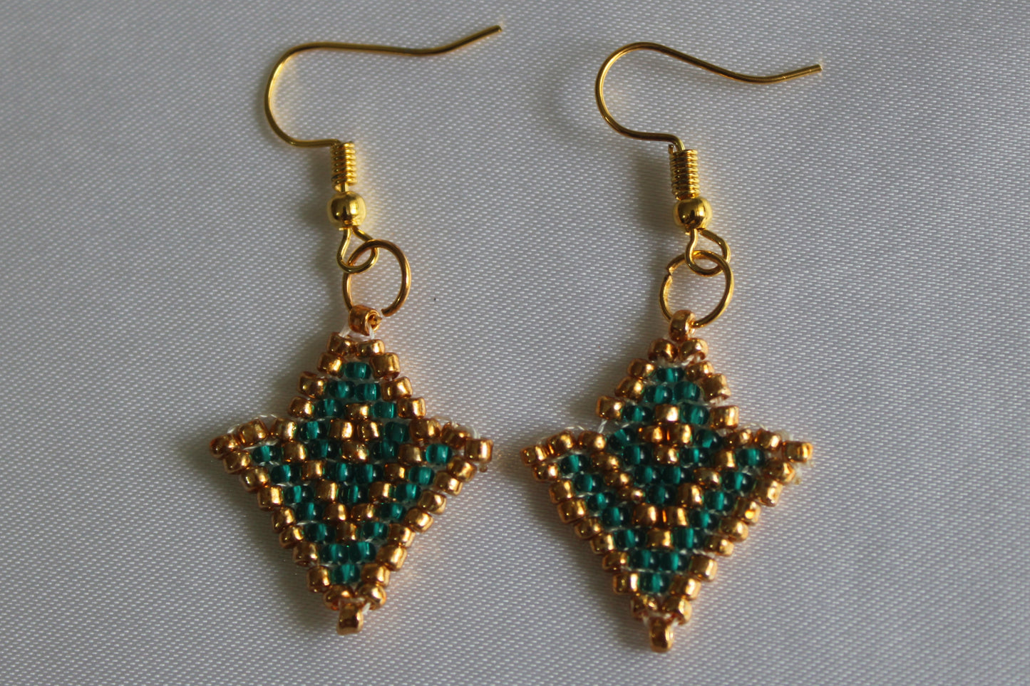A pair of handmade beaded dangle earrings with gold-tone hooks and diamond-shaped charms featuring a green and gold bead pattern, displayed on a light gray fabric background.