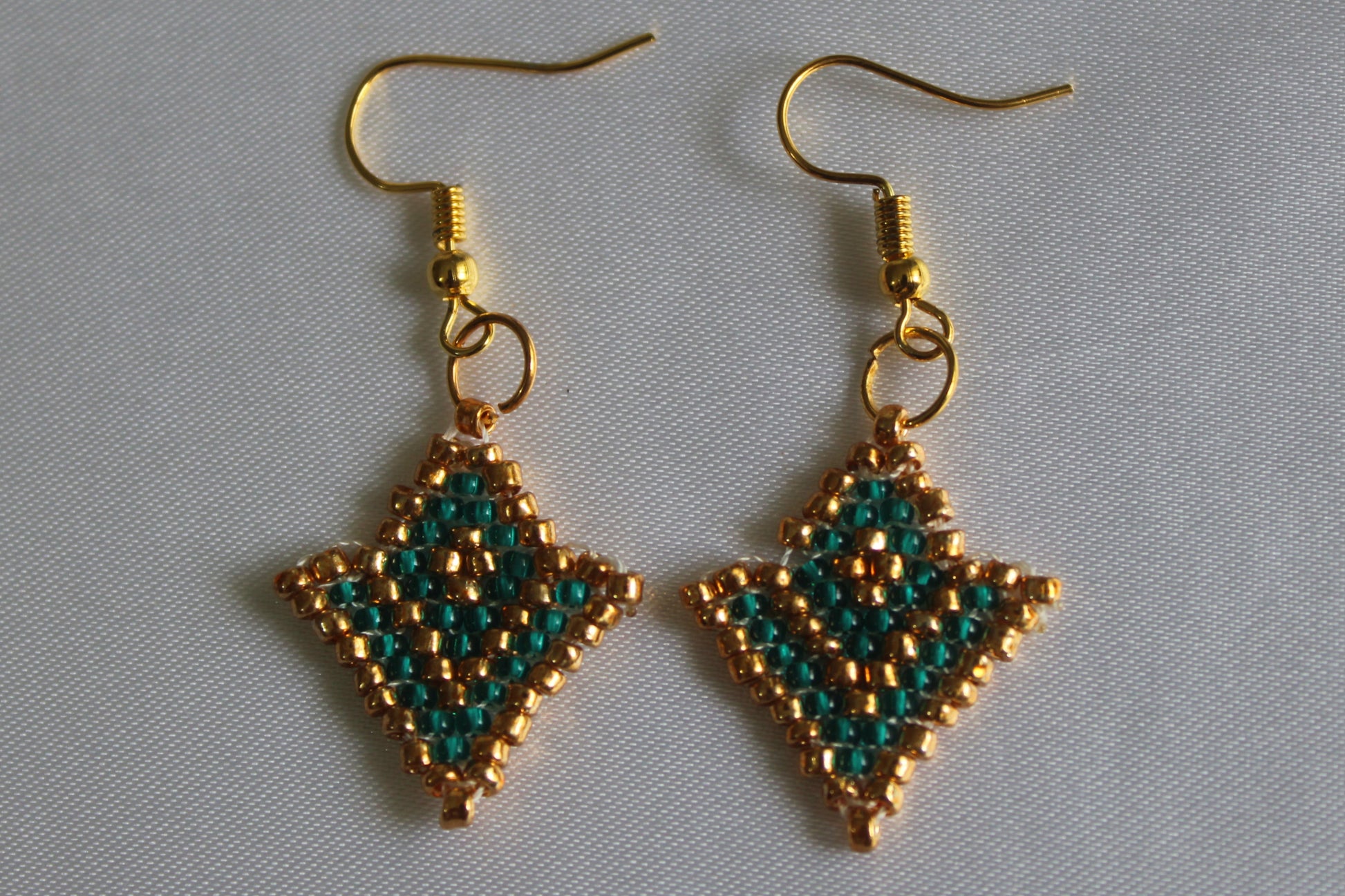 A pair of handmade beaded dangle earrings with gold-tone hooks and diamond-shaped charms featuring a green and gold bead pattern, displayed on a light gray fabric background.