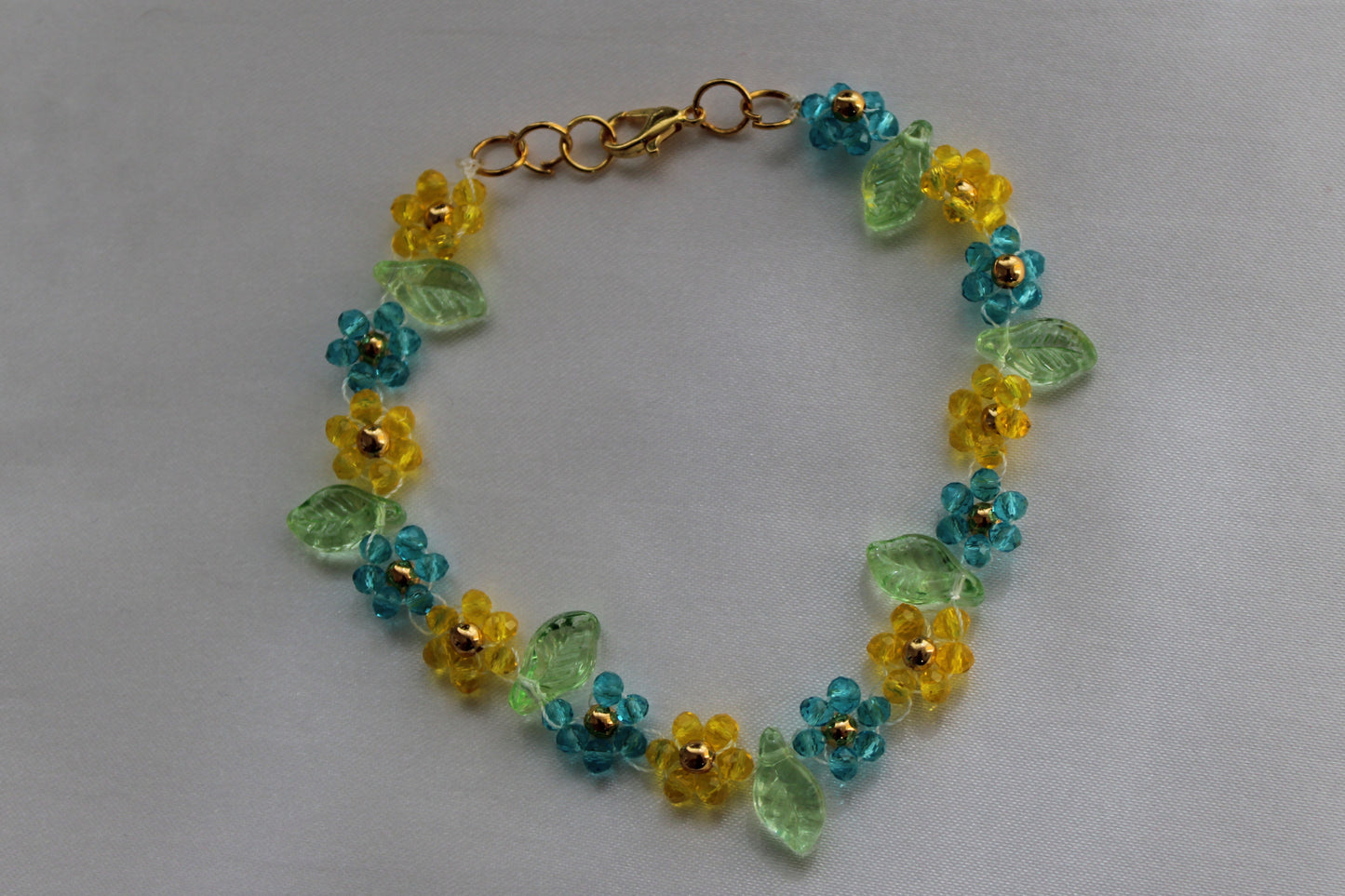 A handmade beaded bracelet featuring yellow flower beads with gold centers, turquoise blue flower beads, and translucent light green leaves, finished with a gold clasp, shown on a neutral light gray background.