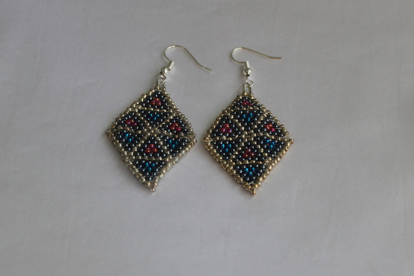 Handmade diamond-shaped geometric beaded earrings featuring black, metallic blue, silver, and red seed beads with fishhook ear wires on a white background.