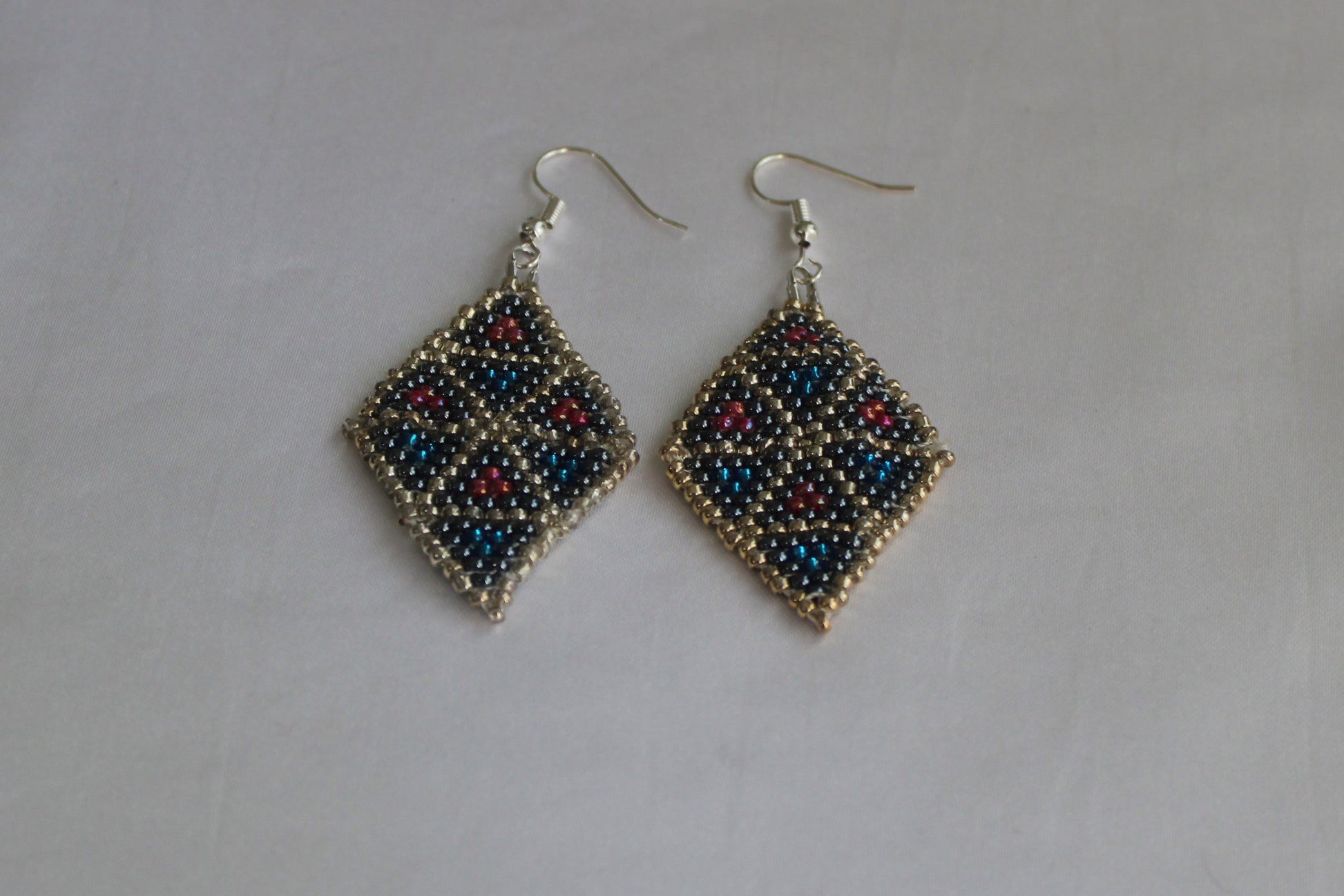Handmade diamond-shaped geometric beaded earrings featuring black, metallic blue, silver, and red seed beads with fishhook ear wires on a white background.