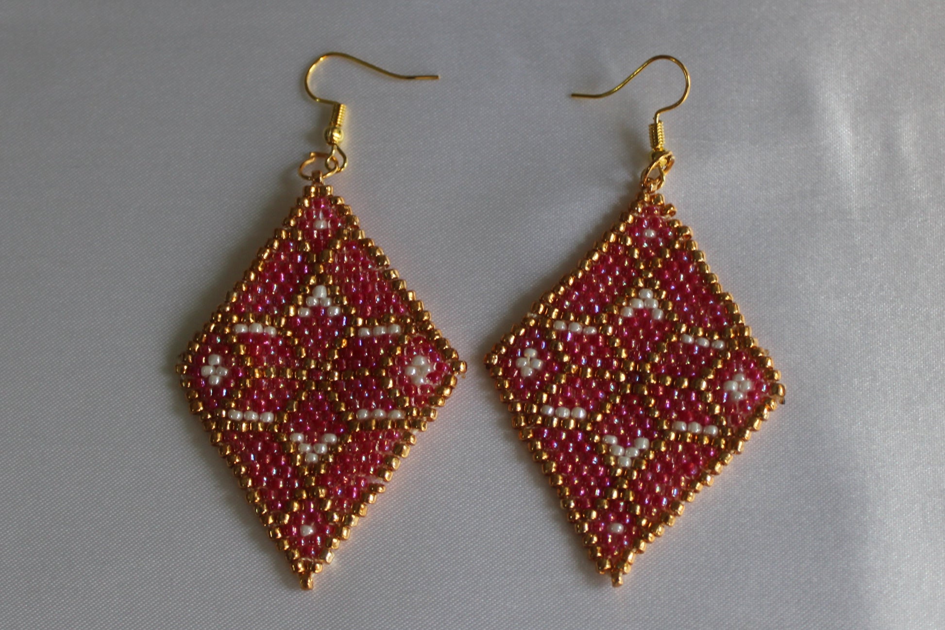 A pair of handmade beaded dangle earrings with gold fishhook wires. The diamond-shaped beads feature a geometric pattern in red, gold, and white, set against a plain light gray background.