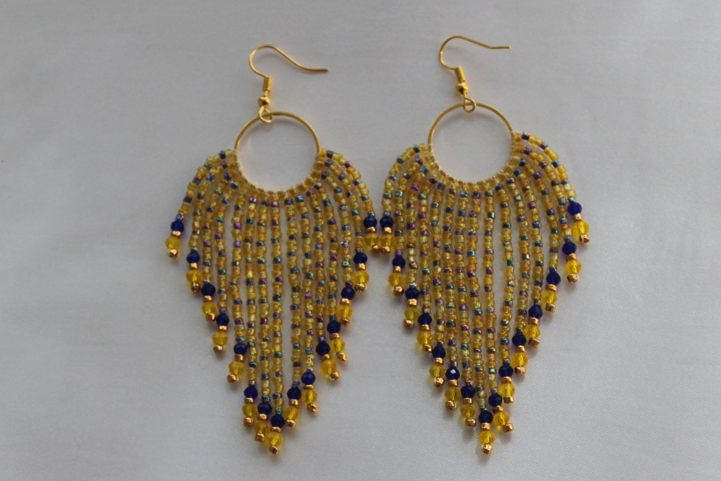 A pair of gold-tone dangle earrings with circular hoops and cascading fringe beads in yellow, blue, and gold, displayed on a light gray background.