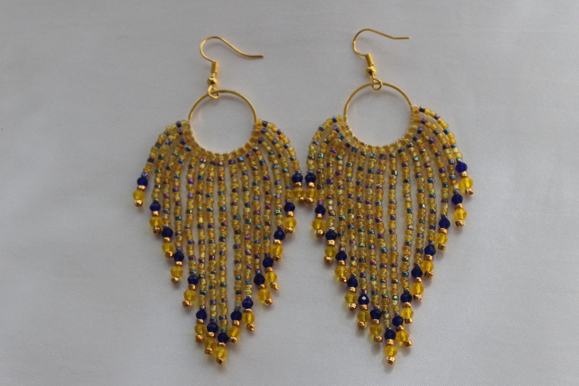 A pair of gold-tone dangle earrings with circular hoops and cascading fringe beads in yellow, blue, and gold, displayed on a light gray background.