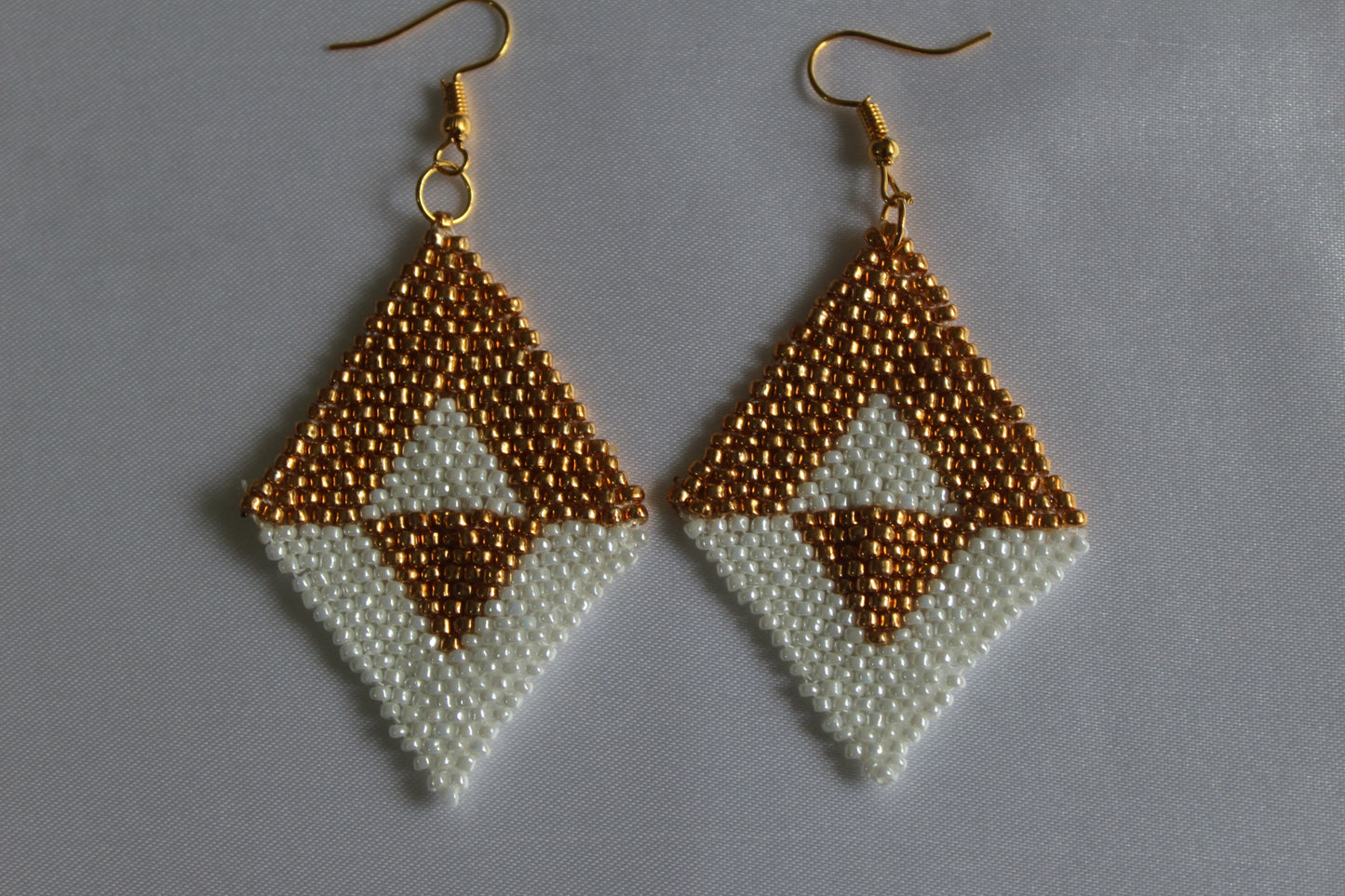 A pair of handmade beaded dangle earrings with gold fishhook wires. The earrings feature a geometric diamond design with a textured gold top section, a white middle section, and a smaller gold triangle at the bottom, all made from small seed beads.