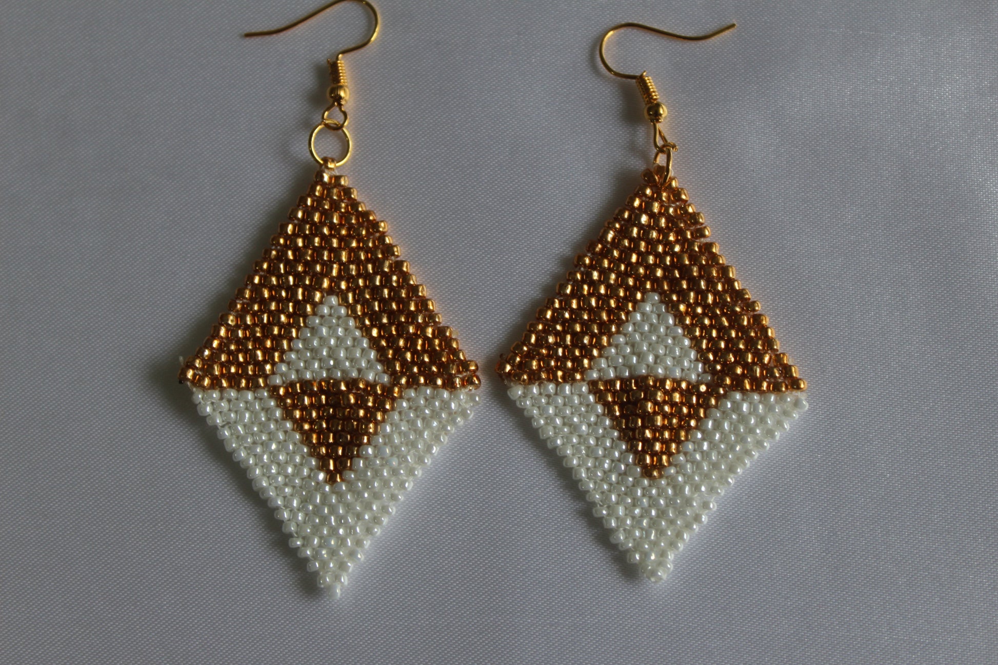 A pair of handmade beaded dangle earrings with gold fishhook wires. The earrings feature a geometric diamond design with a textured gold top section, a white middle section, and a smaller gold triangle at the bottom, all made from small seed beads.