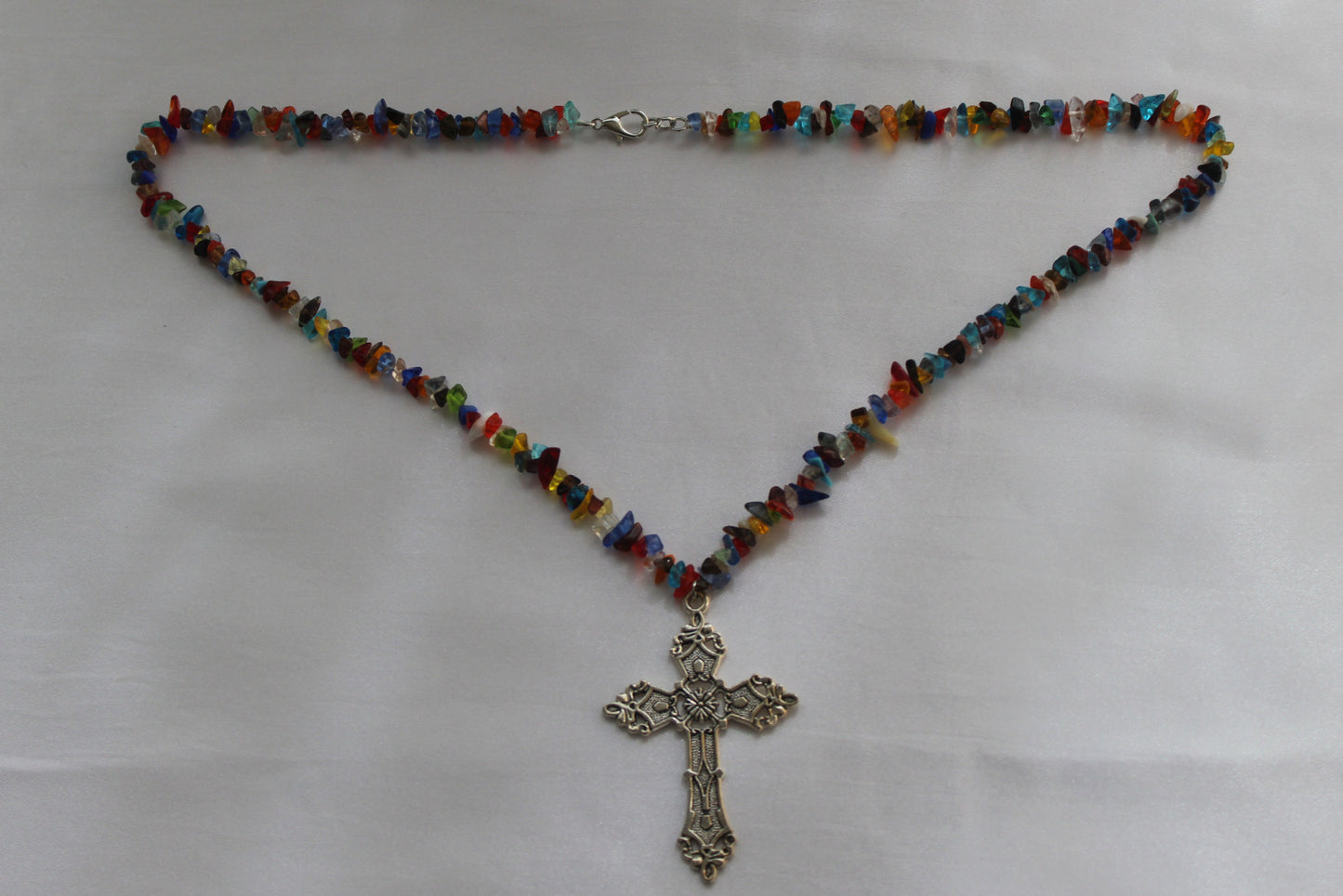 A beaded necklace with small multicolored glass chips and a silver-toned ornate cross pendant, laid flat on a white fabric background.
