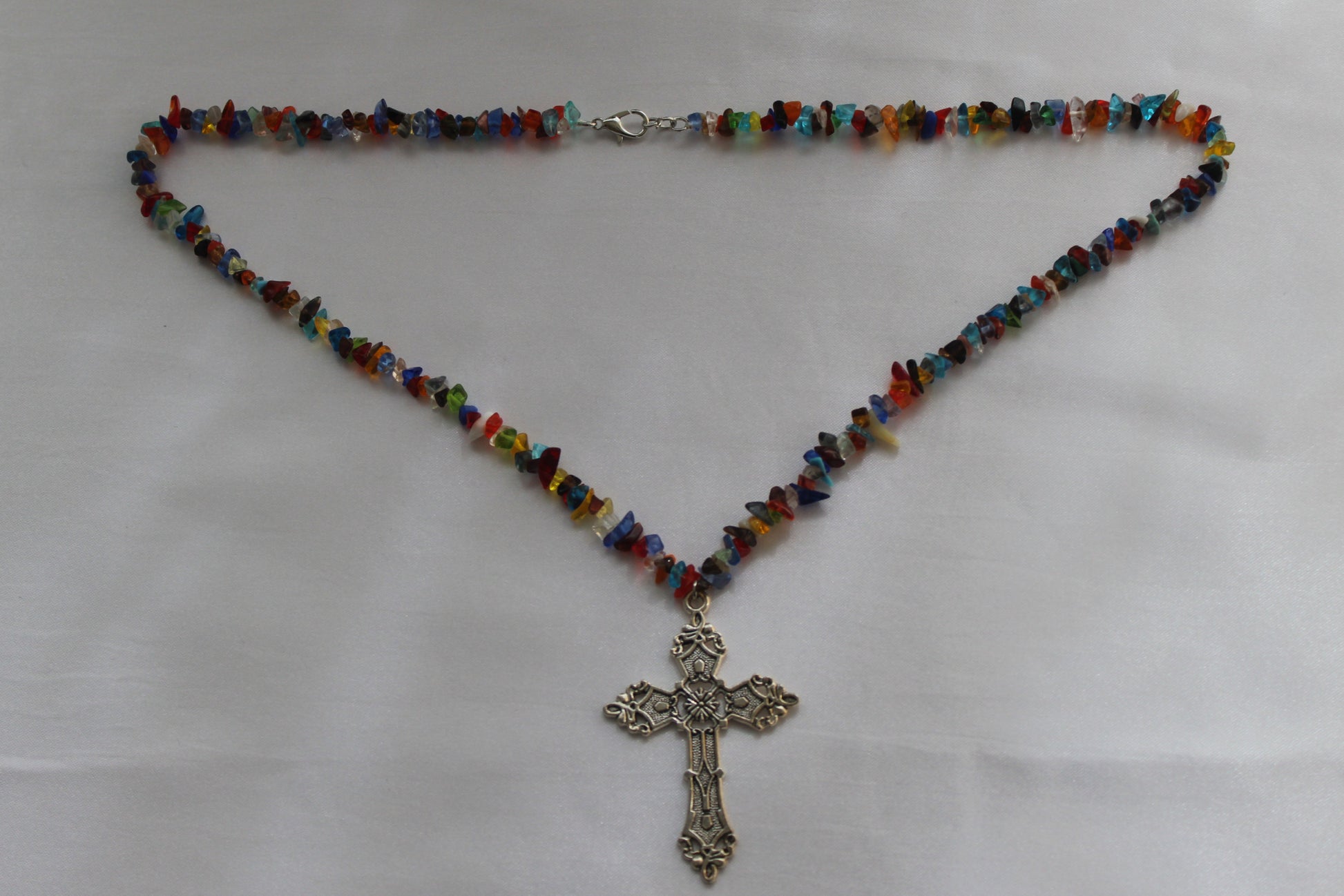 A beaded necklace with small multicolored glass chips and a silver-toned ornate cross pendant, laid flat on a white fabric background.
