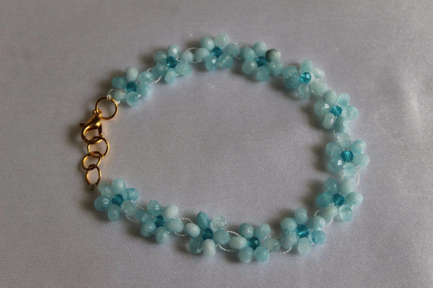 A beaded flower bracelet made of light blue faceted beads with a gold clasp, displayed on a light gray fabric background.