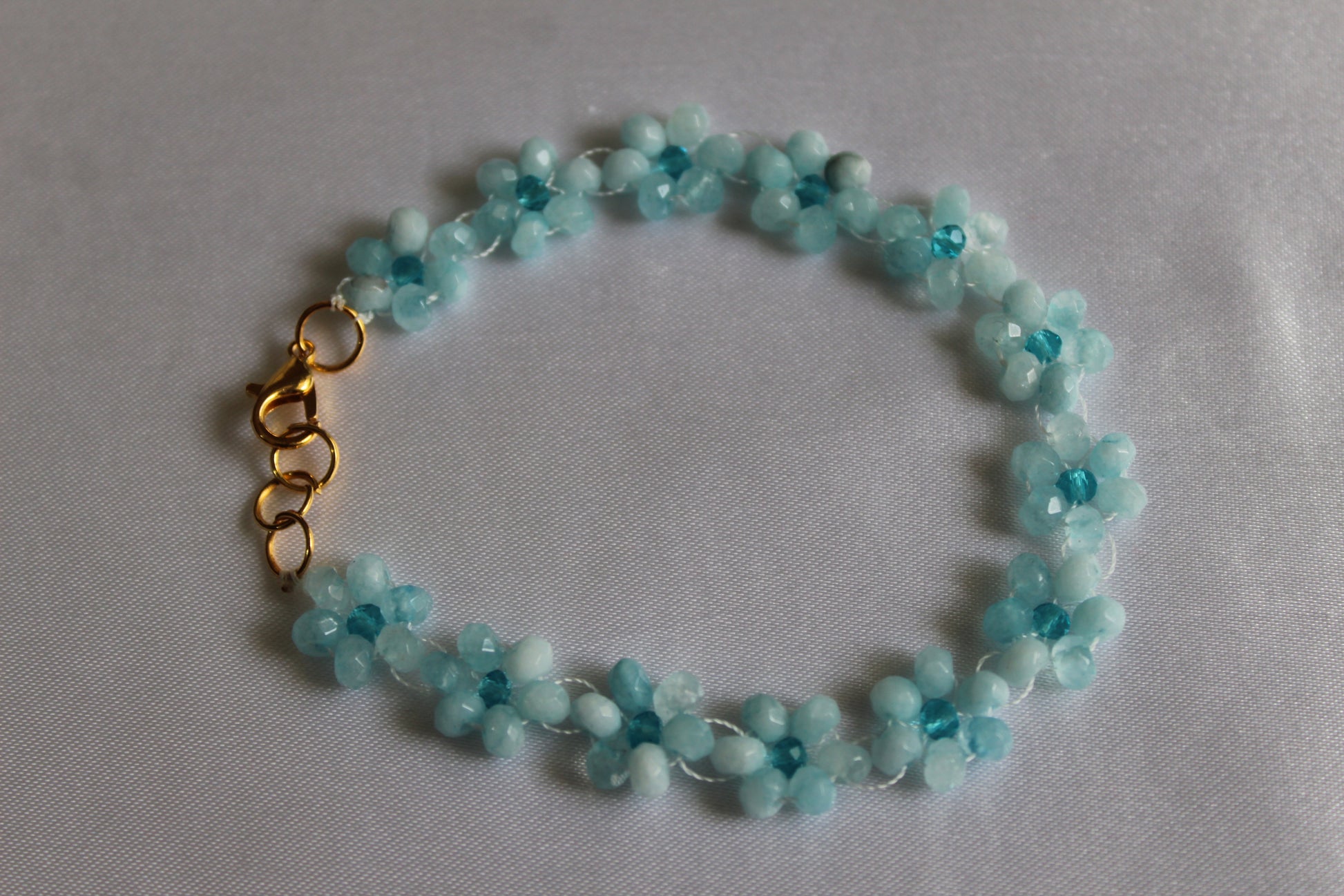 A beaded flower bracelet made of light blue faceted beads with a gold clasp, displayed on a light gray fabric background.