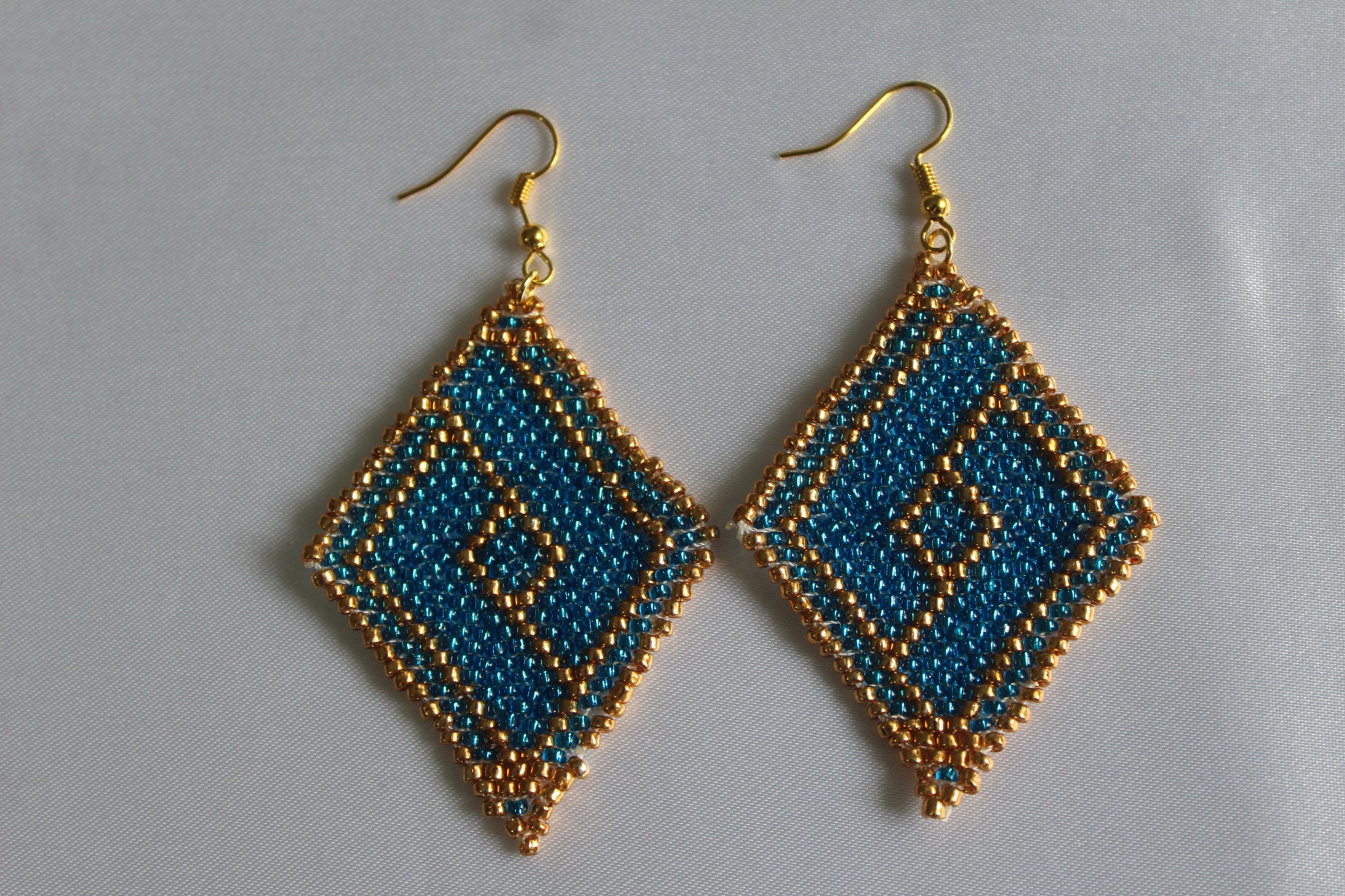 Handmade teal blue and gold diamond-shaped seed bead earrings featuring an intricate geometric pattern with gold-tone fish hook ear wires, lightweight boho statement jewellery.