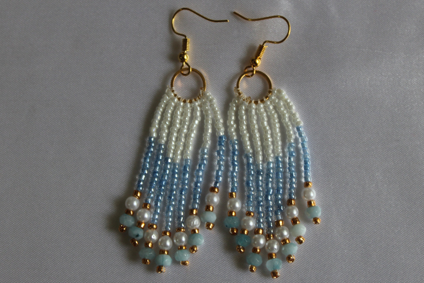 A pair of handmade dangle earrings with gold fishhook wires, circular gold rings, and cascading beaded fringe in white, light blue, and pale aqua, finished with small gold beads.