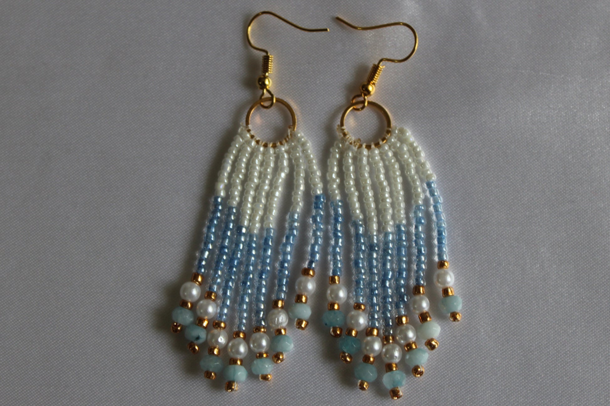A pair of handmade dangle earrings with gold fishhook wires, circular gold rings, and cascading beaded fringe in white, light blue, and pale aqua, finished with small gold beads.