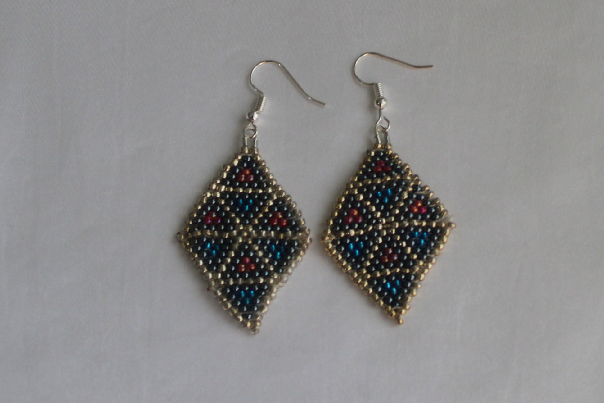 Handmade diamond-shaped geometric beaded earrings featuring black, metallic blue, silver, and red seed beads with fishhook ear wires on a white background.